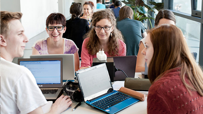 Studierende sitzen in einem Student Space des Fakultätsgebäudes mit ihren Laptops und unterhalten sich.