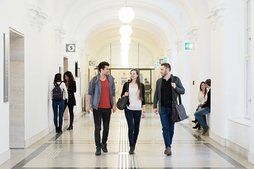 Drei Studierende gehen einen Gang im historischen Hauptgebäude der Universität Wien entlang und unterhalten sich. Im Hintergrund sind weitere Gruppen von Studierenden zu sehen.